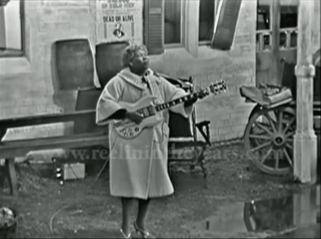 Black-and-white of Sister Rosetta Tharpe playing guitar on old-timey set.